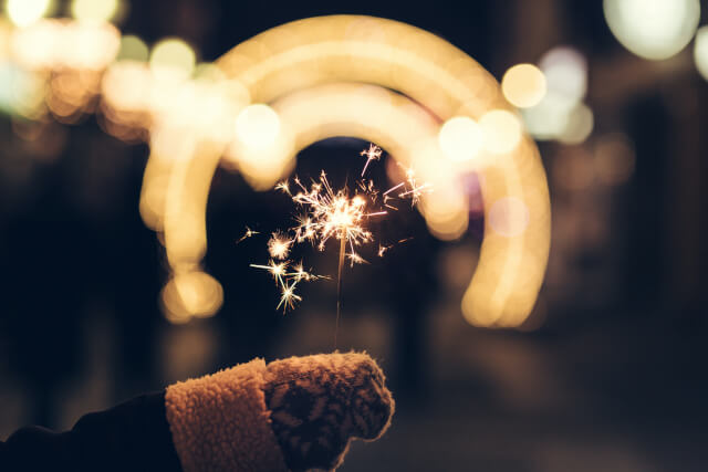 A hand holding a lit sparkler with a blurred light in the distance